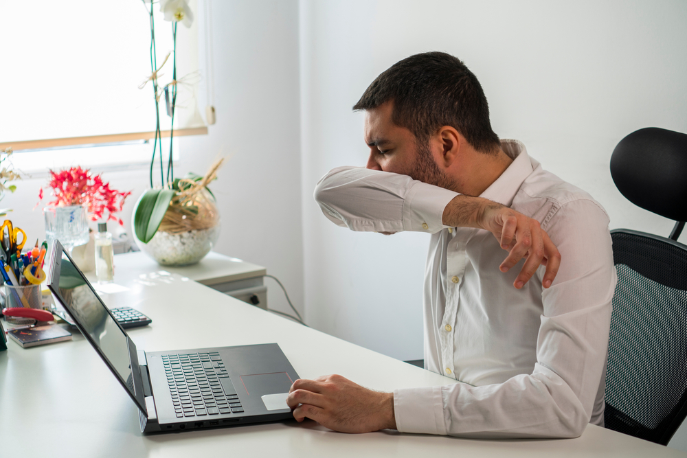 Man sneezing into his shirt sleeve while working on laptop